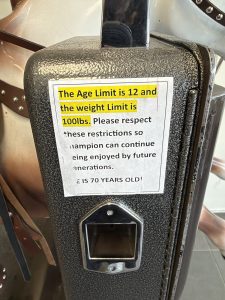 The sign printed and taped to the side of the coin box, which reads as such:
The Age Limit is 12 and the weight limit is 100 lbs, please respect these restrictions so Champion can continue being enjoyed by future generations. SHE IS 70 YEARS OLD!
The first sentence is highlighted in yellow.